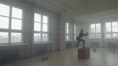 Sıkı spor giyim giymiş, dayanıklılık egzersizi yapan, dumanlı çatı katı tarzı spor salonu stüdyosunda tek başına boks antrenmanına atlayan genç bir kadının ağır çekimde tam çekimi.