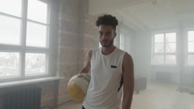 Ağır tıp topunu kolunun altında tutan kararlı Afro-Amerikan sporcunun orta boy portresi dumanlı çatı katı tarzı eğitim odasının penceresinin yanında duruyor.