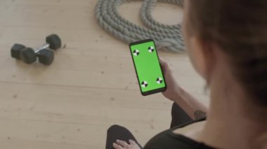 Siyah spor kıyafetli, kapalı alanda spor yaparken akıllı telefon kullanan tanınmayan bir kadının omuz üstünden çekimi.