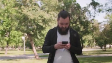 Güneşli bir günde parkta yürüyen ve cep telefonunu kullanan sakallı bir gencin fotoğrafını çekiyorum.