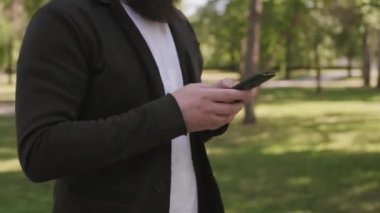 Sakallı bir gencin parkta yürürken ve cep telefonuyla yazarken elde çekilen görüntüler.