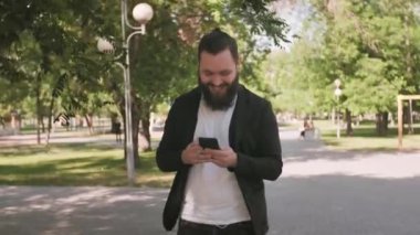 Sakallı, mutlu bir gencin görüntüsü cep telefonuyla konuşuyor ve güneşli bir günde parkta yürüyor.