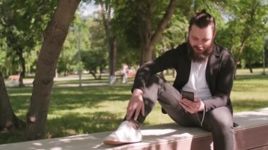 Güneşli bir günde parktaki uzun sırtüstü bankta oturan ve cep telefonuyla video konuşması yapan sakallı genç adamın PAN görüntüsü.