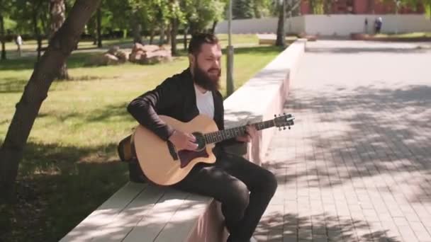 在阳光灿烂的日子里 追踪坐在公园长椅上 留着胡子 弹吉他的年轻街头男歌手的照片 — 图库视频影像