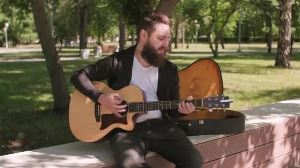 在阳光明媚的夏日 一个留着胡子的年轻人坐在公园的长椅上弹奏吉它 — 图库视频影像