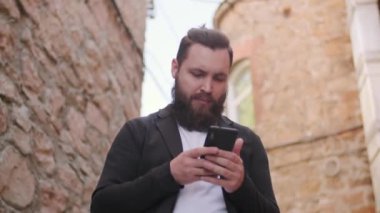 Sakallı genç adamın düşük açılı görüntüsü. Dışarıda duruyor ve cep telefonu kullanıyor.