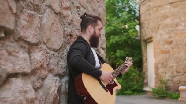 跟踪一个留着胡子的年轻人站在狭窄街道上的旧建筑旁弹奏吉他的镜头 — 图库视频影像