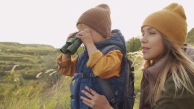 5 yaşındaki meraklı bir çocuğun dürbünle baktığı ve yürüyüş sırasında tepenin manzarasının keyfini çıkardığı yavaş çekim. Neşeli genç annesi bir şeyi işaret edip onunla konuşuyor.