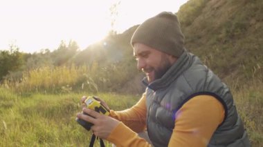 Şiş yelek ve bereli mutlu sakallı adamın güneşli bir günde gülümseyen ve fotoğraf çeken fotoğrafının yavaş bir yan görüntüsü.