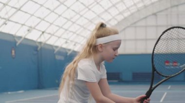 Beyaz tenli küçük bir kızın tenis raketi tutarken, tenis kortunda kameraya bakarken, yavaş çekim portresi.