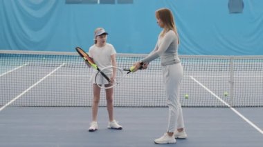 Yavaş çekimde, genç bayan tenis koçu küçük kıza tenis raketi eğitimini kapalı alanda birlikte nasıl atacağını gösteriyor.