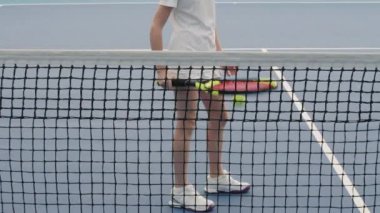 Tenis kortunda tek başına duran küçük kızın yavaş çekimde çekilmiş portresi tenis raketiyle topa vuruyor sonra da kameraya bakıp gülümsüyor.