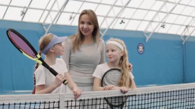 Tenis antrenmanı sırasında kameraya gülümseyen iki küçük kız arasında duran genç bayan koçun orta yavaş portresi.