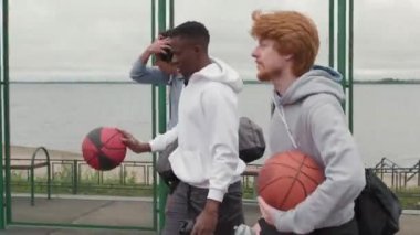 Spor çantaları, su şişeleri ve basketbol topları taşıyan kapüşonlu üç genç adamın kasvetli bir günde açık hava basketbol sahasında yürüdüğünü gösteren bir fotoğraf.