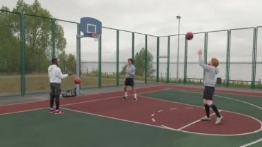 Kasvetli bir günde dışarıda basketbol sahasında basketbol oynayan üç gencin el kamerasıyla çekilmiş görüntüleri.