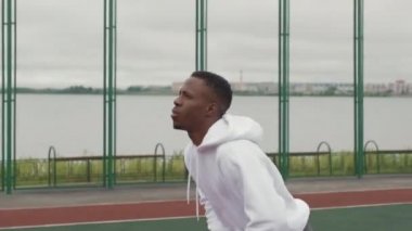Kapüşonlu siyahi adamın kasvetli bir günde basketbol sahasında basketbol oynarken orta boy fotoğrafı.