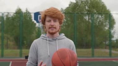 Kızıl saçlı, sakallı, basketbol topu tutan ve dışarıdaki sahada kameraya gülümseyen mutlu bir gencin portre çekimi.