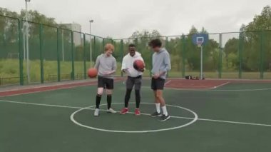 Dışarıda basketbol sahasında takılan ve kameraya gülümseyen üç adamın el kamerasıyla çekilmiş görüntüsü.