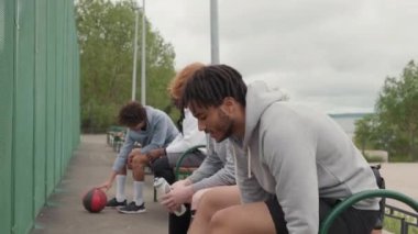 Kasvetli bir günde arkadaşça sokak topu oynamadan önce dışarıda basketbol sahasının yanındaki banklarda oturan ve ayakkabı bağcıklarını bağlayan neşeli gençlerin el kamerasıyla çekilmiş bir fotoğrafı.