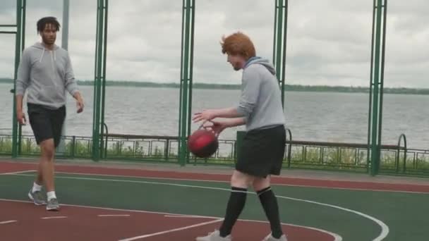 フリーバスケットボールコートで友人とストリートボールをプレイしながら男投げボールと得点の遅い追跡ショット — ストック動画