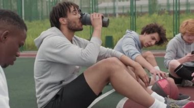 Bir grup gencin açık hava basketbol sahasında oturup maçtan sonra dinlenmelerini izlemek. Telefonlarını ve içme sularını kontrol ediyorlar.