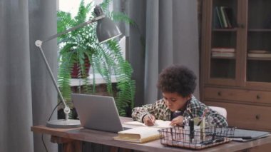 8 yaşındaki akıllı ve tatlı afro çocuğun orta ölçekli görüntüsü oturma odasında dizüstü bilgisayarla çalışıyor, defter ile yazıyor.