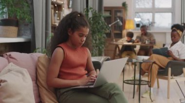 Annesi koltukta oturup kitap okurken ve babası arka planda oğluyla ödev yaparken, 10 yaşında akıllı afro bir kızın laptopta oturup ders çalışmasının orta ölçekli resmi.