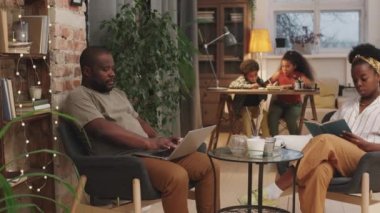 Dört kişilik Afro-Amerikan ailesinin sıcak oturma odasında geçirdiği orta boy PAN fotoğrafı. Dizüstü bilgisayarda çalışan bir adam ve karısı kitap okurken, kardeşler arka planda ödev yapıyor.