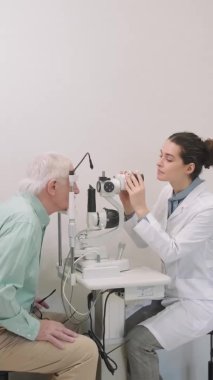 Göz hekimliği bürosundaki masada oturan yaşlı bir adamın normal göz muayenesi yaparken çekilmiş dikey bir fotoğrafı.