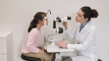 Modern klinikteki göz doktoruna düzenli göz kontrolü yapan küçük bir kızın yan görüntüsü. Kadın doktor, küçük kızın görme yeteneğini fosfor makinesiyle test ediyor.
