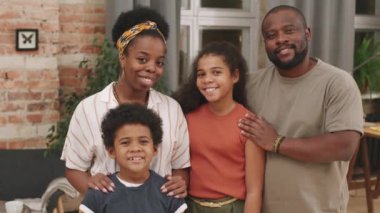 Dört kişilik mutlu Afro-Amerikan ailesinin orta boy portresi. Oturma odasında kameraya gülümsüyor.