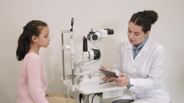 在现代诊所 中等比例的小女孩回答女眼科医生关于预约医生的问题 在谘询期间在数码平板电脑上作笔记的妇女 — 图库视频影像