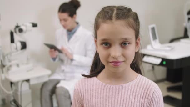 中特写Pan肖像 镜头中 一个年轻貌美的小女孩笑着站在眼科医生办公室前拍照 而女医生则坐在工作场所 背景是数码平板电脑 — 图库视频影像