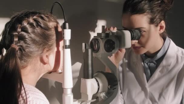 女性眼科専門家の中間閉鎖行う目の検査のために女の子見ますスルーフォトワーマシン座っています日光満たされた医師のオフィス — ストック動画