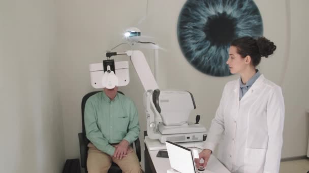 若い女性眼科医の中出し眼科用アイチャートプロジェクターを使用しながらシニア男行います目の検査で現代オフィス — ストック動画