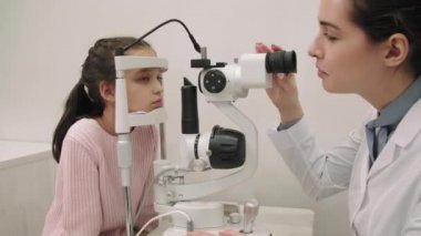 Kafkasyalı küçük bir kızın modern klinikte gözünün phoropter makinesiyle test edildiği orta boy bir fotoğraf. Bayan göz doktoru küçük bir kız için göz kontrolü yapıyor.