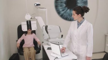 Küçük kız ve bayan göz doktorunun modern optik ekipmanlarla klinikte düzenli göz kontrolü yaparken orta boy fotoğrafı.