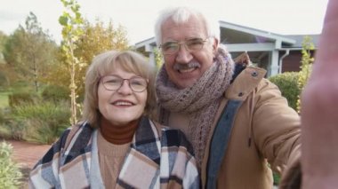 Orta yaşlı, beyaz bir kadın ve kocasının evlerinin yanında dikilirken ve ellerinde cihazlı adamla kameralı görüşme yaparken çekilmiş.