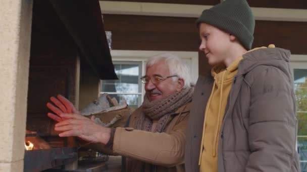 10代の白人少年の中で 暖炉の外に立つ暖かい服を着て 明るい祖父が手を暖めるのを火で見ている — ストック動画