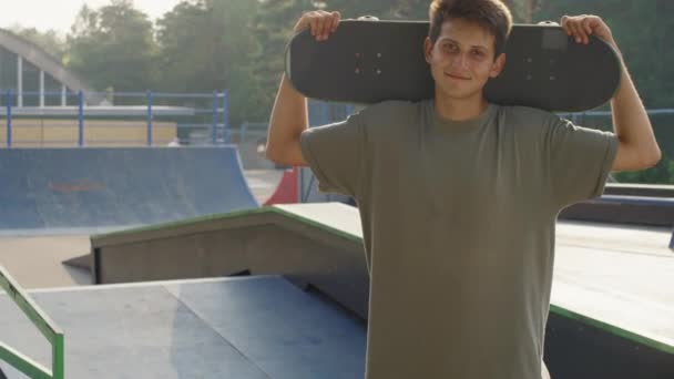 ポートレートは ランプ付きスケートパークでカメラのために笑顔スケートボードと陽気な若い男のショットを傾けます — ストック動画
