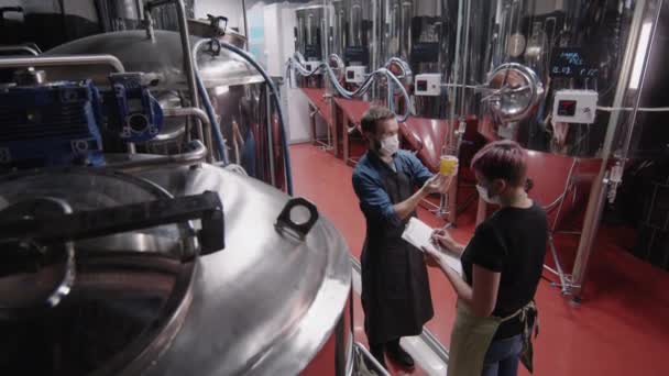 带保护面罩的两名酿酒厂工人用慢镜头拍摄的长角形中镜头 用新生产的饮料检查玻璃杯中的啤酒质量 并在平板电脑上做笔记 — 图库视频影像