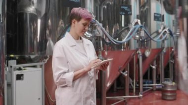 Beyaz laboratuvar önlüklü kadın bira fabrikasında dikilip dijital tablet üzerinde çalışırken kameraya bakıp gülümseyen kadın bira fabrikası müdürünün yavaş çekimleri.