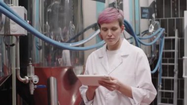 Beyaz önlüklü kadın bira fabrikası müdürünün bira üretim fabrikasında yürüdüğü, bira ekipmanlarını kontrol ettiği dijital tabletle notlar aldığı orta boy bir fotoğraf.