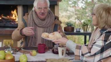 Beyaz eşlerin sıcak kıyafetler giyip açık terasta oturup yemek yedikleri orta boy bir fotoğraf. Kadın kocasına tabaktan turta almasında yardım ediyor.