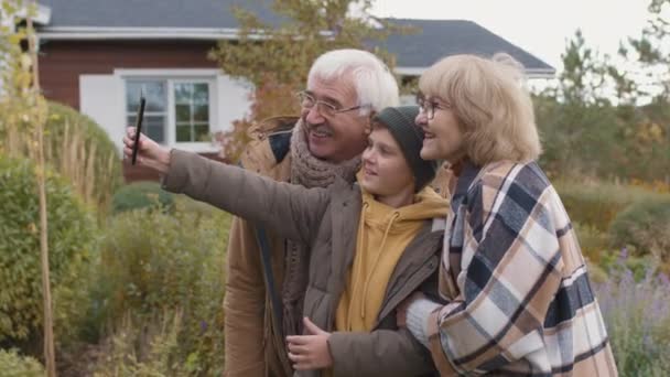 中等比例的白人妇女 男人和他们的小孙子穿着温暖的休闲服站在自家附近的花园里 用电话摄像头自拍 — 图库视频影像