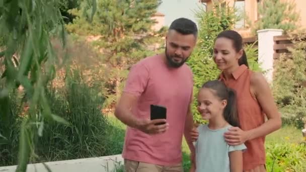 晴れた夏の日に裏庭に立っている3人の陽気な家族の中のショットと父親はスマートフォンで自撮り — ストック動画