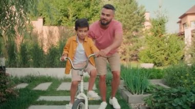 Modern yetişkin bir adamın 8 yaşındaki neşeli oğluna güneşli bir yaz gününde arka bahçede bisiklet sürmeyi öğretirken çekilen yavaş çekim.