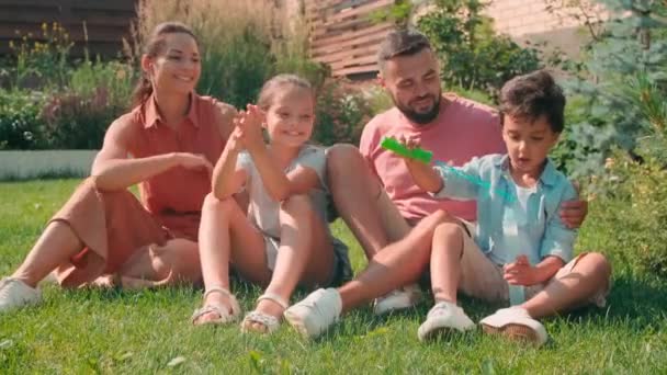 晴れた夏の日に一緒に楽しい4人の幸せな若い家族の完全なショット石鹸泡を作り 裏庭の緑の芝生に座って笑っています — ストック動画