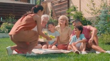 Genç bir kadının taze ev yapımı limonata getirirken çekilmiş düşük açılı fotoğrafı. Bu sırada kocası, çocukları ve büyük ebeveynleri güneşli bir günde bahçede piknik yaparken çimenlerde oturup sohbet ediyorlar.