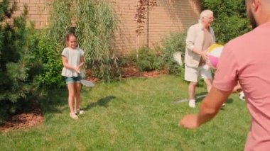 Babasıyla badminton oynayan 10 yaşındaki neşeli bir kızın orta ölçekli çekimi. Bu arada dedesi ve ninesi yaz günü çimenlikte birlikte vakit geçiriyorlar.